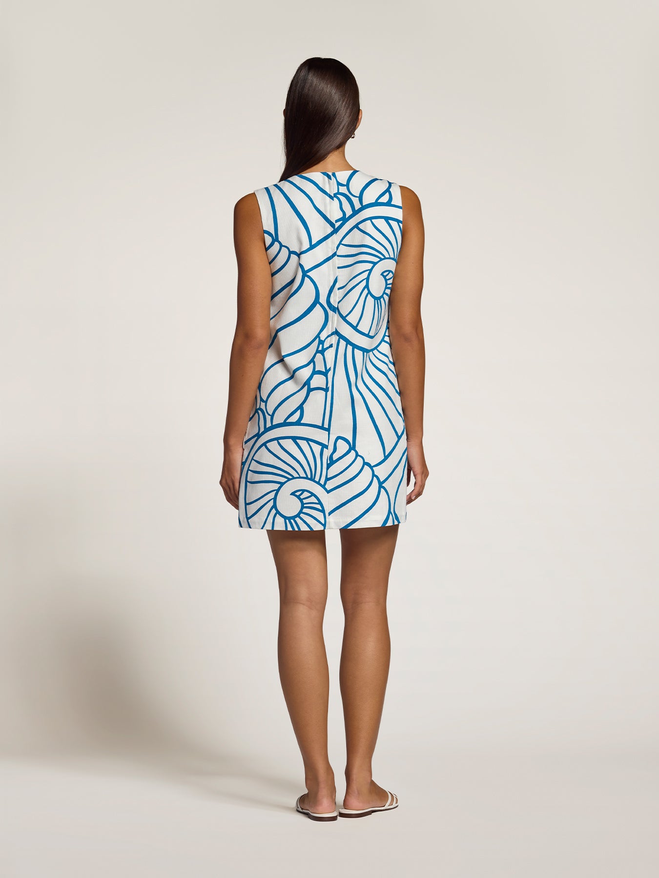 A woman viewed from the back wearing a sleeveless mini dress with a blue and white abstract spiral pattern, standing against a plain light beige studio background.