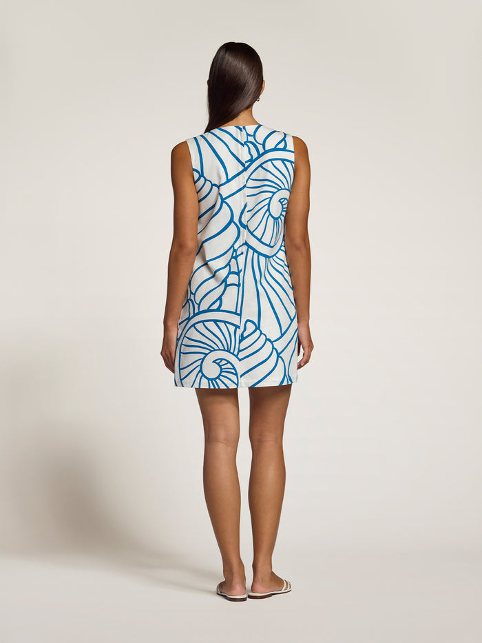 A woman viewed from the back wearing a sleeveless mini dress with a blue and white abstract spiral pattern, standing against a plain light beige studio background.