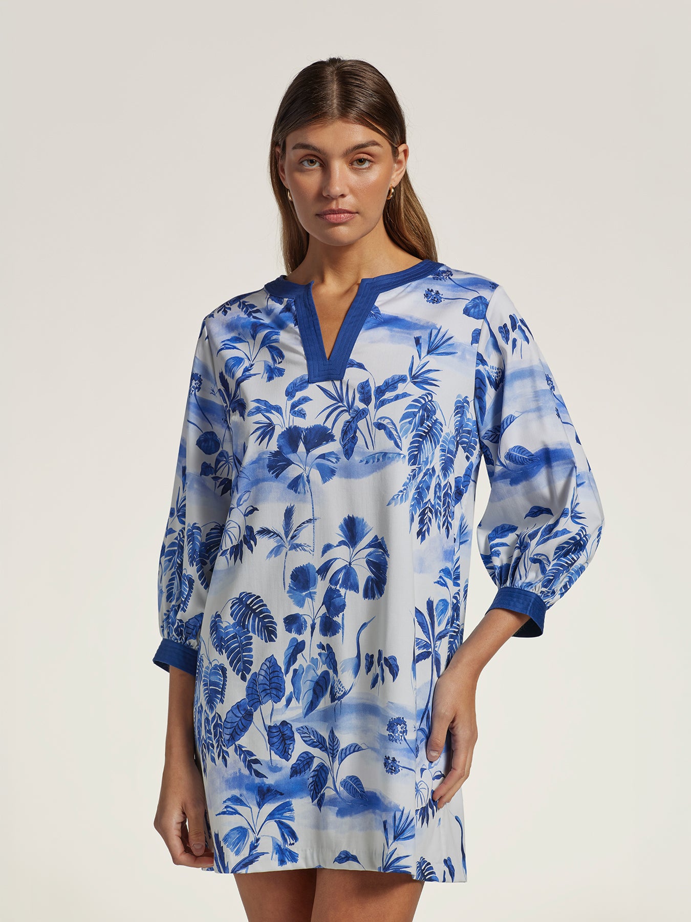 A woman modeling a blue and white tropical leaf print tunic dress with a split neckline and elbow-length sleeves, photographed against a plain light beige studio background.