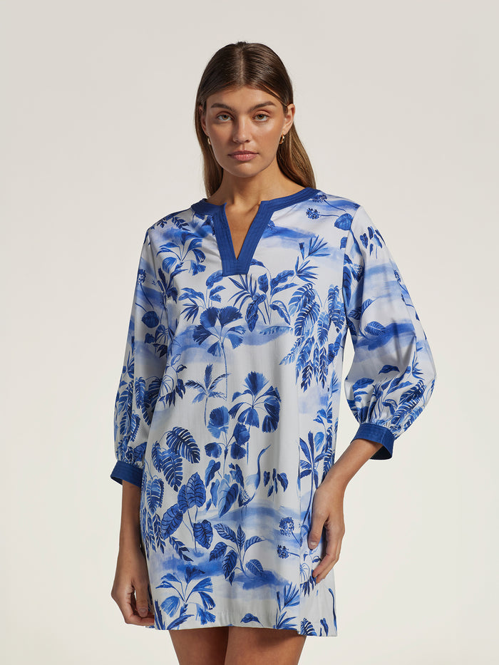 A woman modeling a blue and white tropical leaf print tunic dress with a split neckline and elbow-length sleeves, photographed against a plain light beige studio background.