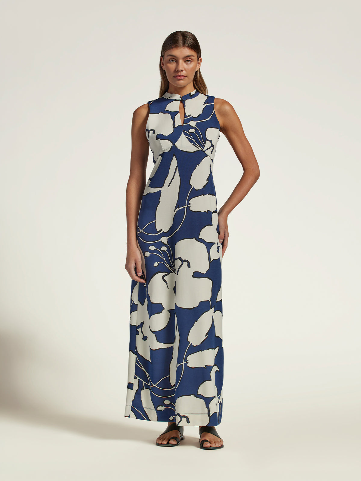 A woman modeling a sleeveless, high-neck maxi dress with a bold navy blue and off-white floral pattern, standing against a plain light beige studio background.