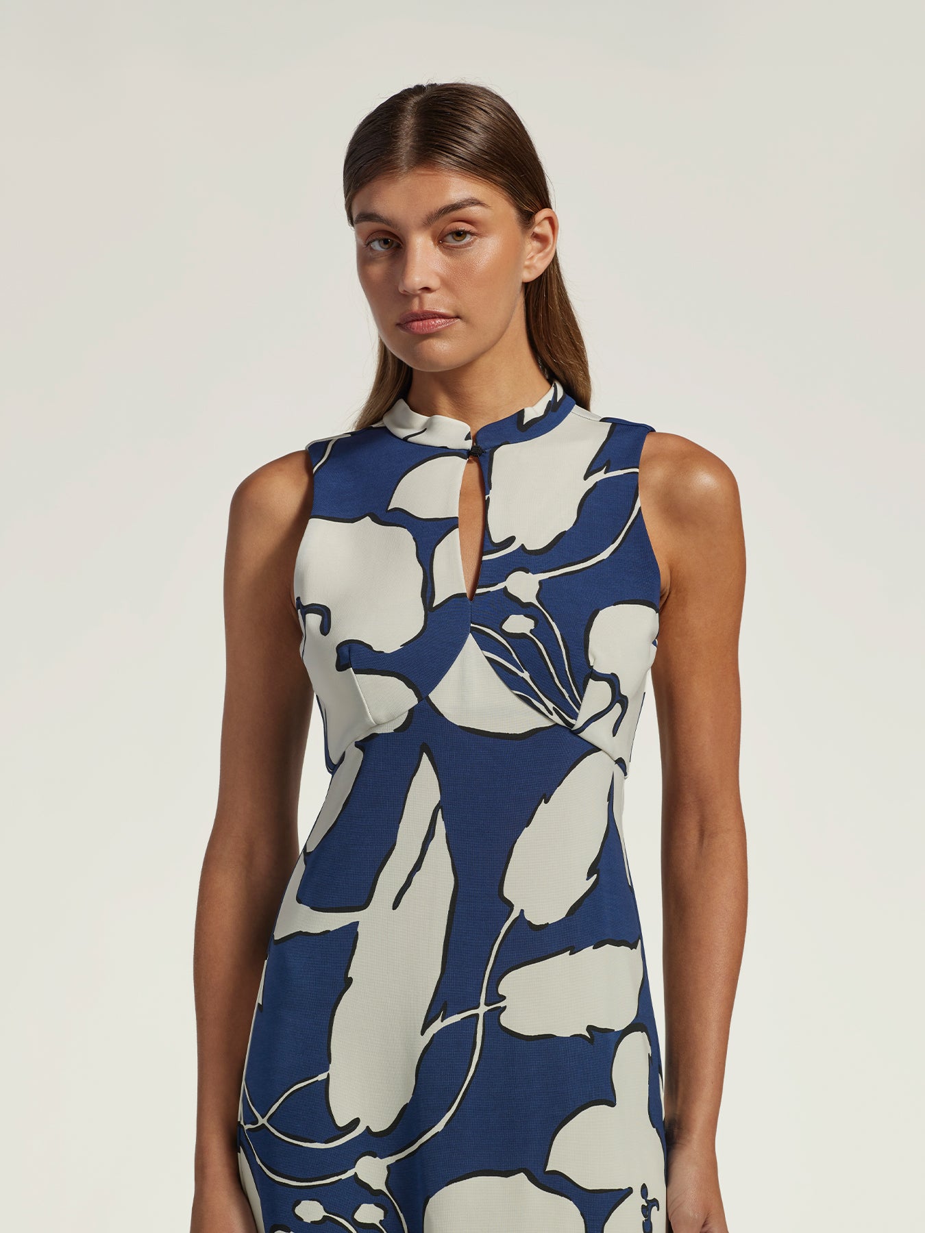 A woman modeling a sleeveless high-neck dress with a keyhole cutout and an abstract floral pattern in navy blue and off-white, set against a plain light gray studio background.