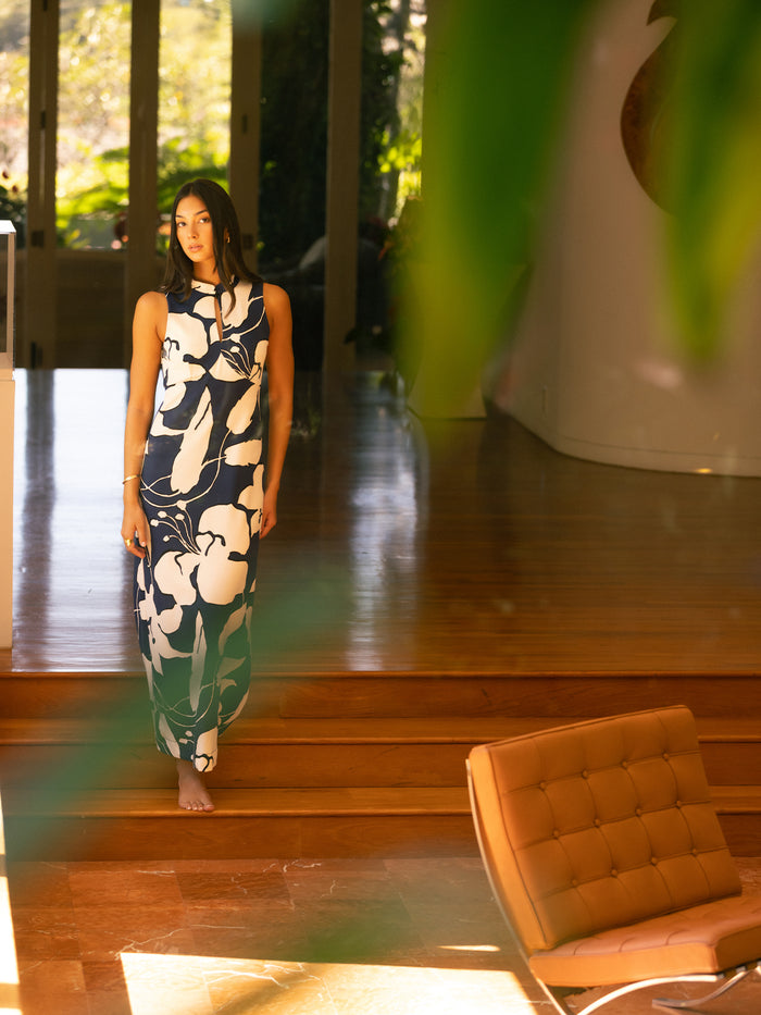 A woman walks down wooden stairs wearing a sleeveless navy maxi dress with a large white floral pattern. The photo is taken in a modern home interior with warm sunlight, glass doors, and a tufted leather chair in the foreground.