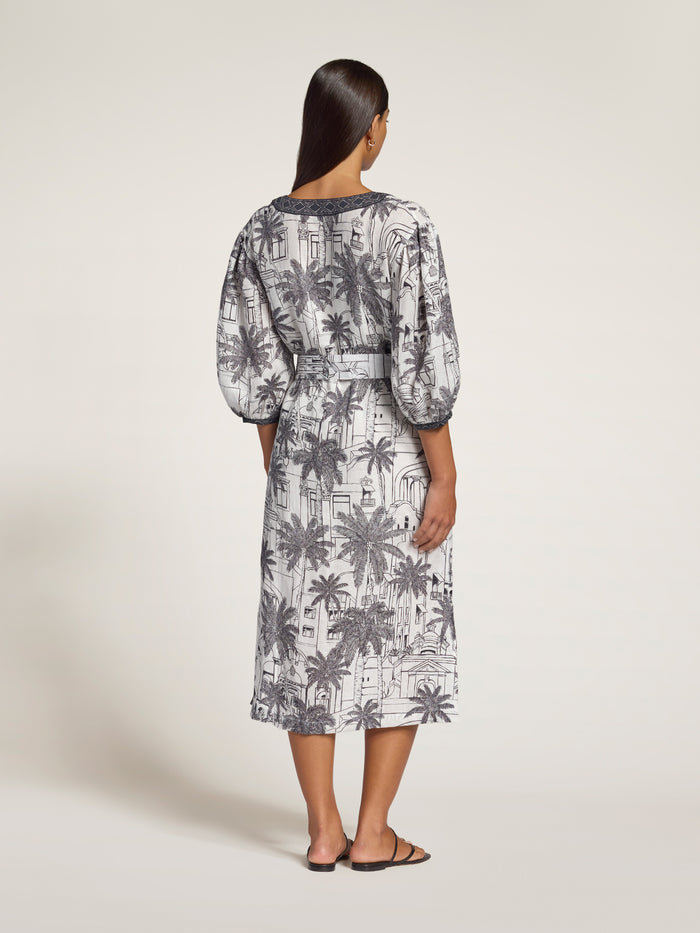 A woman viewed from the back wearing a midi dress with a gray and white palm tree pattern, featuring puff sleeves and a tie at the waist, set against a plain light beige studio background.