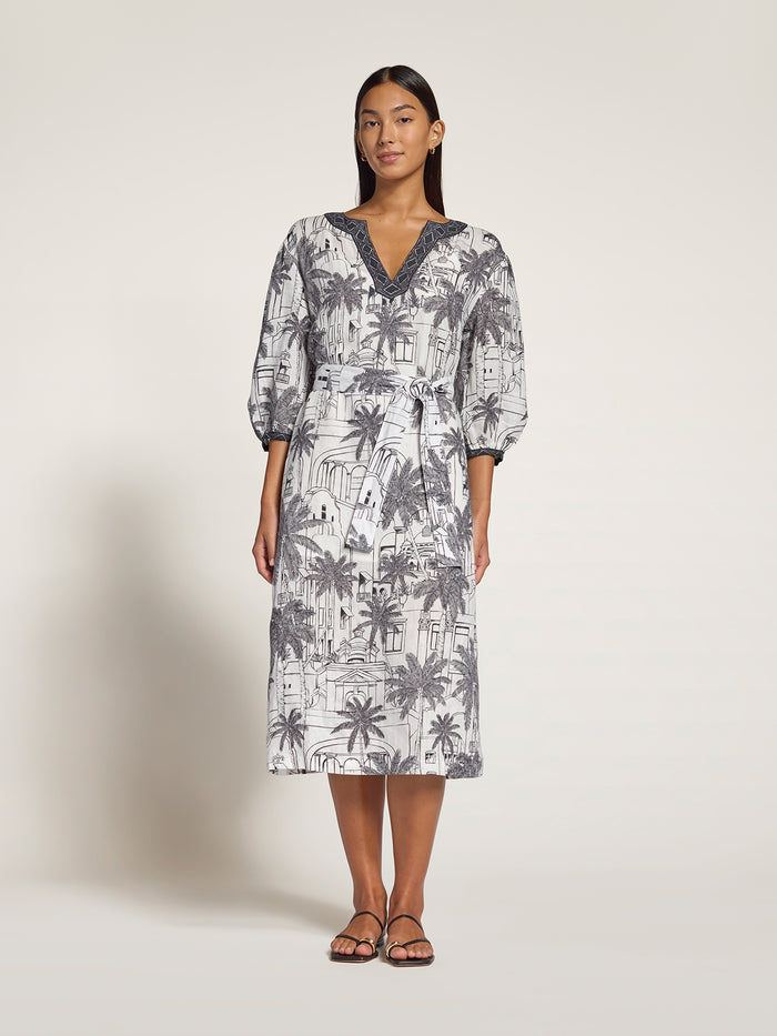 A woman modeling a midi-length dress with a gray and white tropical palm tree print, featuring a V-neckline with dark blue-gray trim and 3/4 puff sleeves, set against a plain light beige studio background.