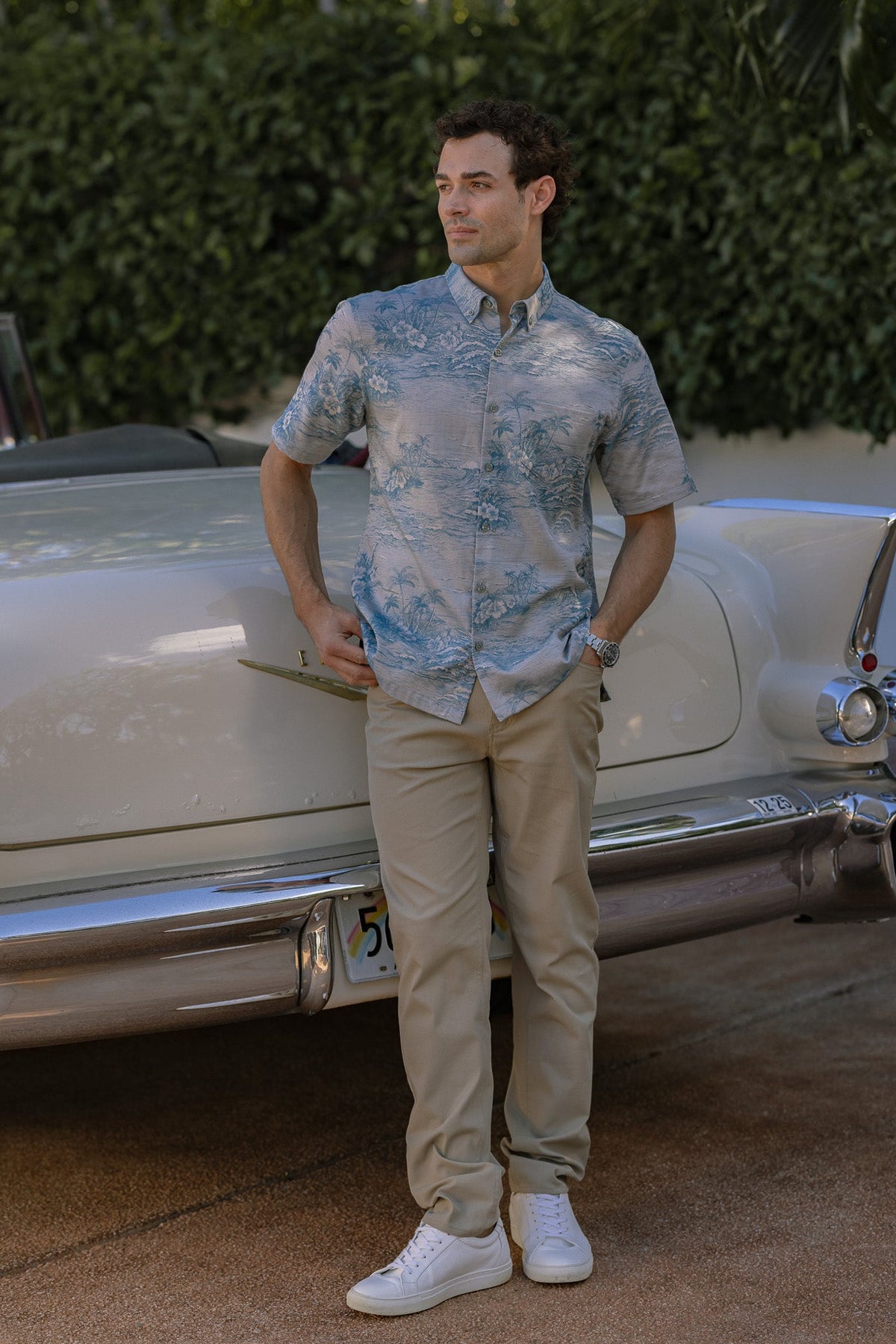 A man in a tropical print button-up shirt, khaki pants, and white sneakers leans against a vintage white car outdoors.