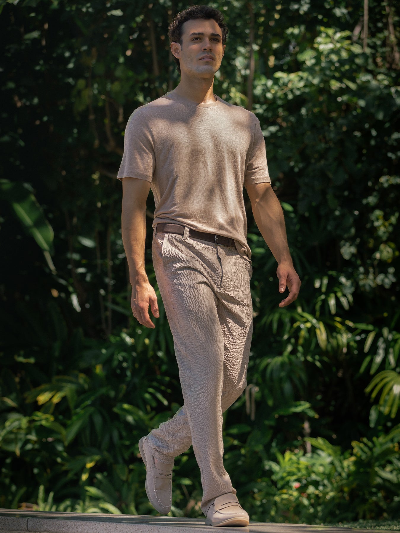 Light beige t-shirt and matching beige trousers, and shoes with a dark belt, set against a lush green background of foliage.