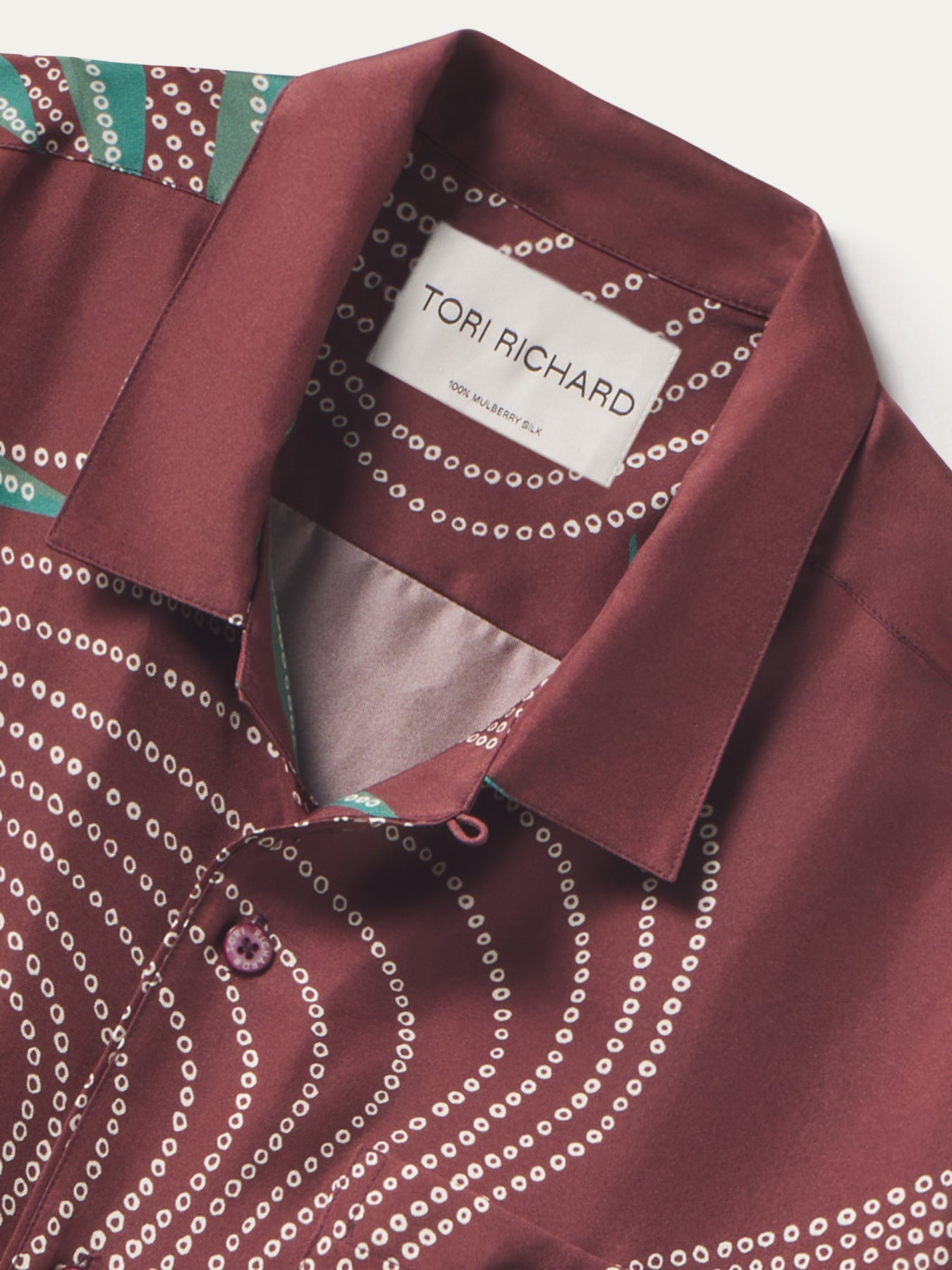 Close-up of a collared button-up shirt by Tori Richard, featuring a deep burgundy base with white dotted concentric circle patterns and a small teal patterned section near the collar.