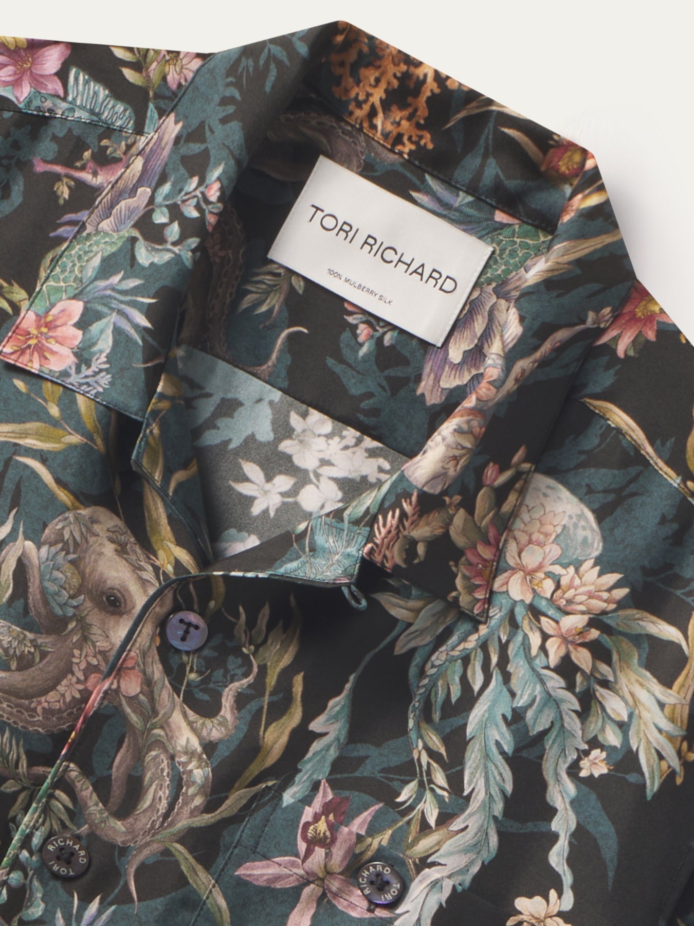 A close-up of a Tori Richard men's button-down shirt featuring a dark green tropical print with octopus, floral, and bird motifs in pink, beige, and teal tones.