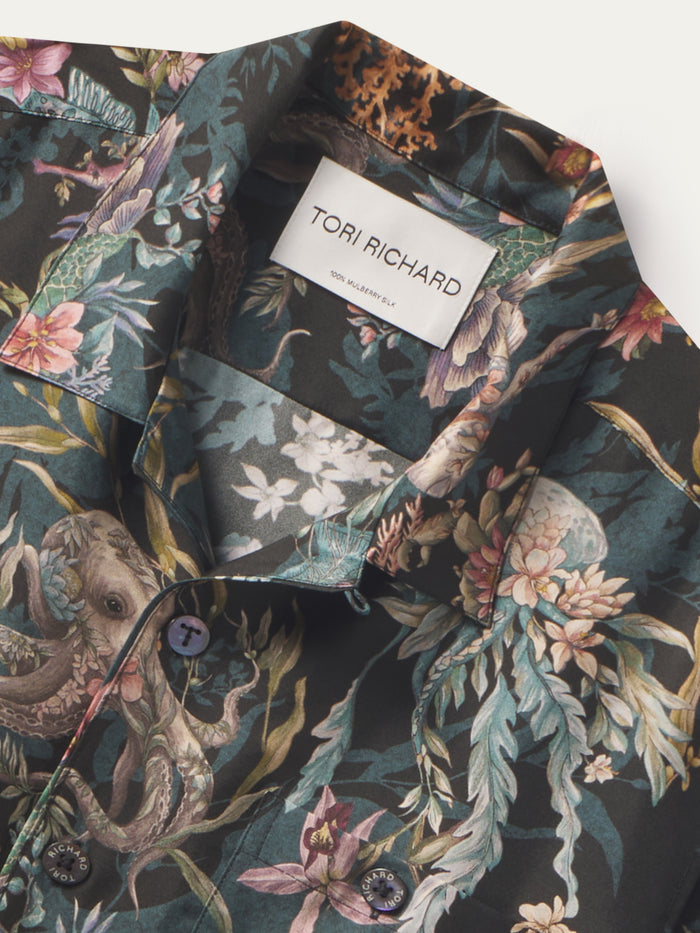 A close-up of a Tori Richard men's button-down shirt featuring a dark green tropical print with octopus, floral, and bird motifs in pink, beige, and teal tones.