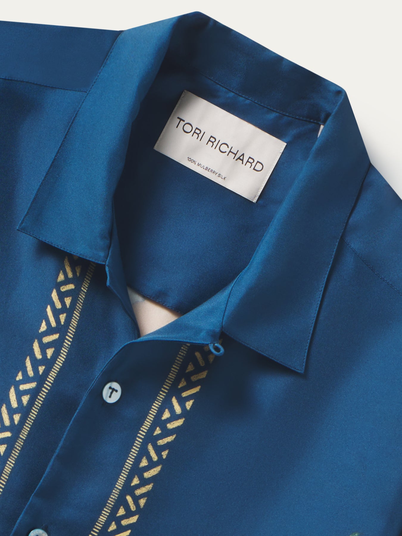 Close-up of a blue collared shirt by Tori Richard, showing the inner neck label and gold geometric embroidery running down the front placket.