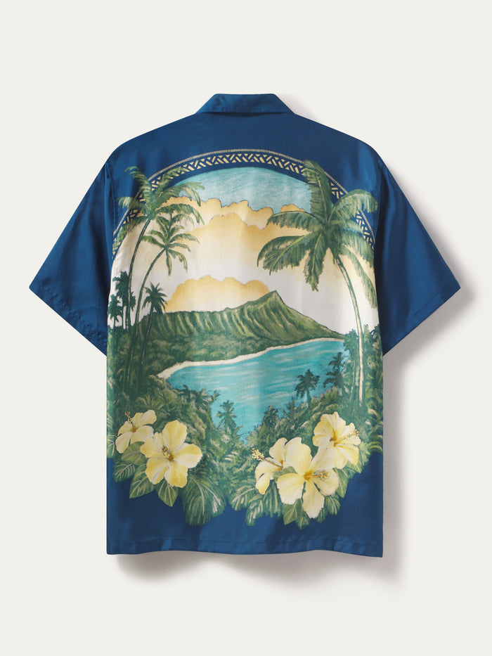 A blue short-sleeve camp shirt shown from the back, featuring a large tropical beach and mountain landscape print with palm trees, hibiscus flowers, and a scenic sunset.