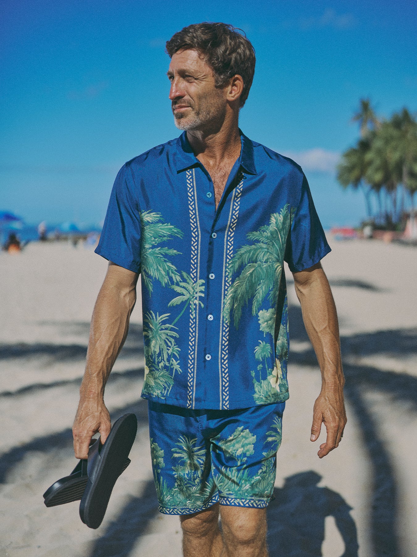 A man walking on a sandy beach wearing a blue short-sleeve button-up shirt and matching shorts with a green palm tree tropical print, holding black sandals.