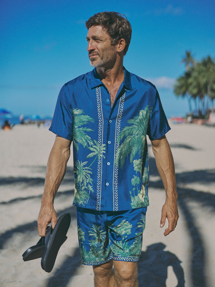 A man walking on a sandy beach wearing a blue short-sleeve button-up shirt and matching shorts with a green palm tree tropical print, holding black sandals.