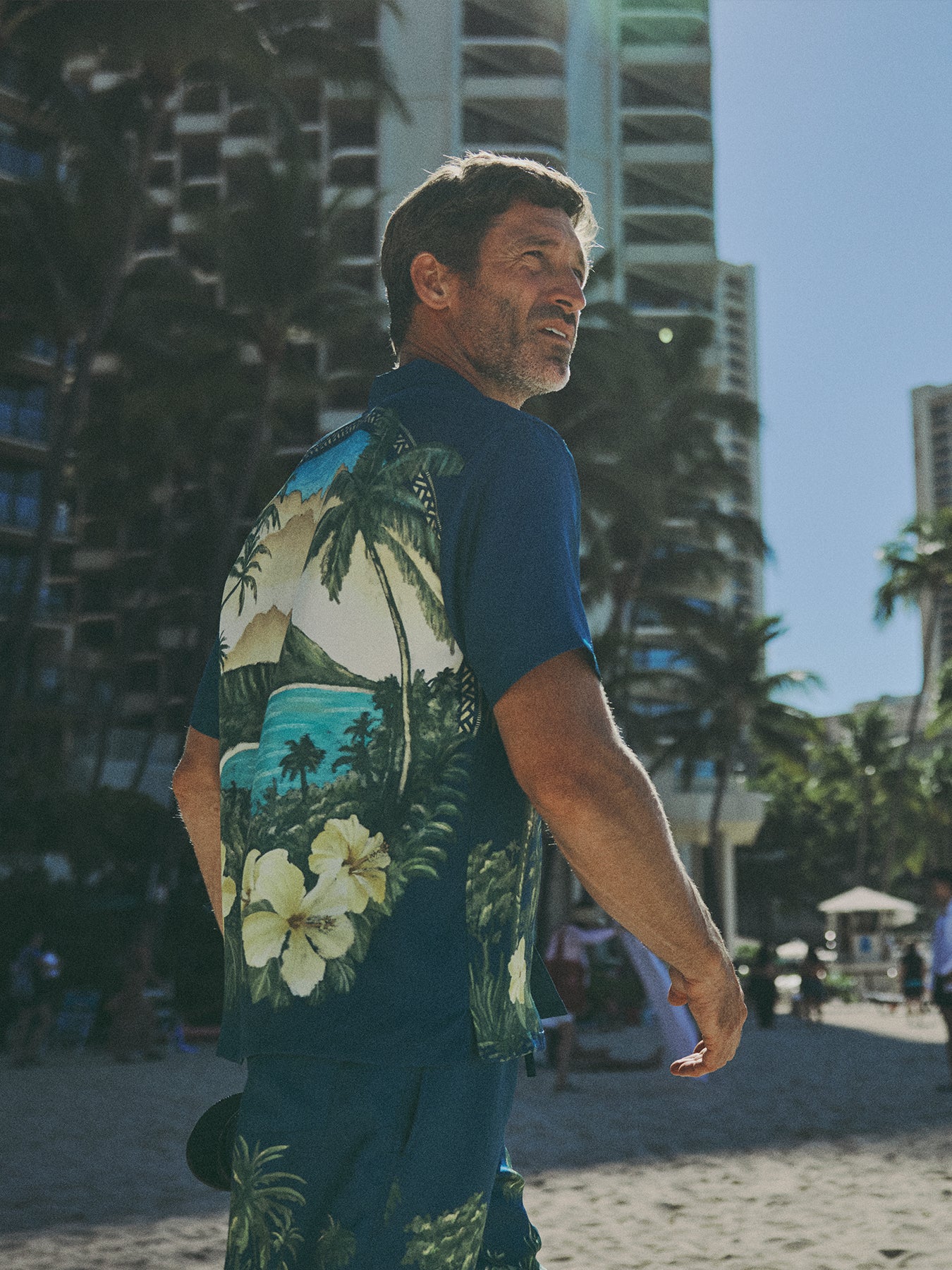 A man wearing a dark blue short-sleeve Hawaiian shirt with a tropical beach print, standing on a sandy beach with palm trees and high-rise buildings in the background.