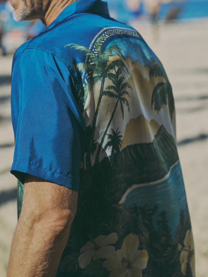A man wearing a blue short-sleeve button-up shirt with a tropical beach print featuring palm trees, mountains, and flowers, photographed outdoors on a sandy beach.