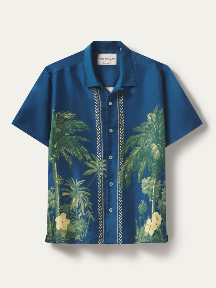 A short-sleeve button-up camp shirt in deep blue with a tropical palm tree and hibiscus flower print, featuring decorative gold patterned trim down the front placket.
