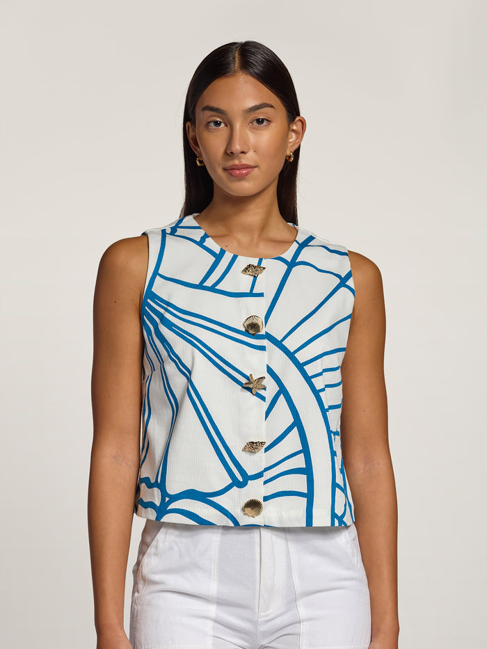 A woman modeling a sleeveless white top with a blue abstract line pattern and decorative gold-tone buttons, paired with white pants, against a plain light gray studio background.