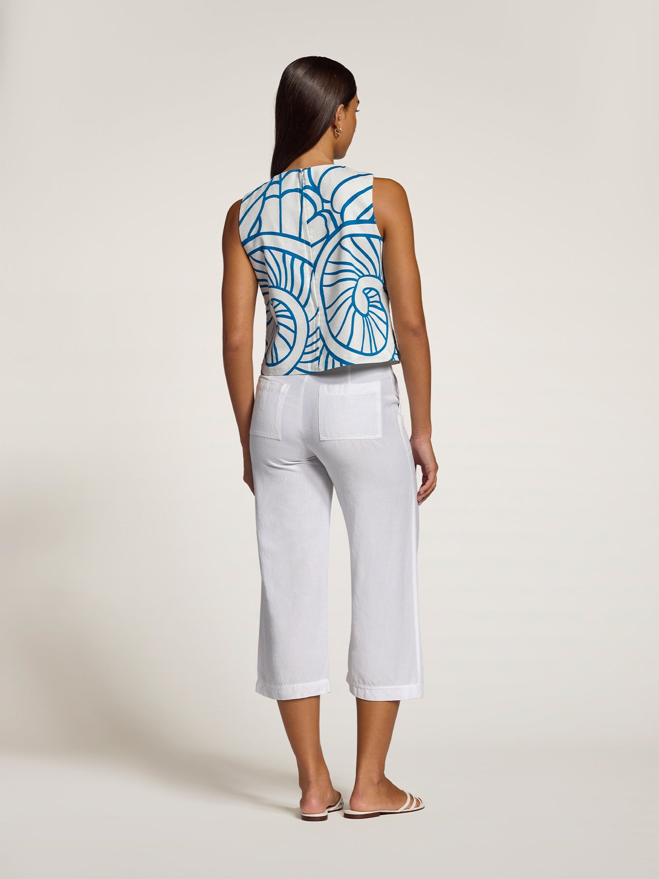 A woman is shown from the back wearing a sleeveless top with a blue and white geometric pattern and white cropped pants, standing against a plain light beige studio background.