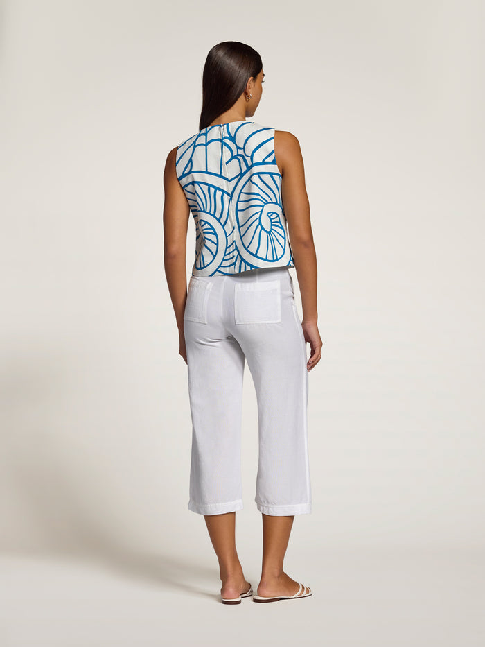 A woman is shown from the back wearing a sleeveless top with a blue and white geometric pattern and white cropped pants, standing against a plain light beige studio background.