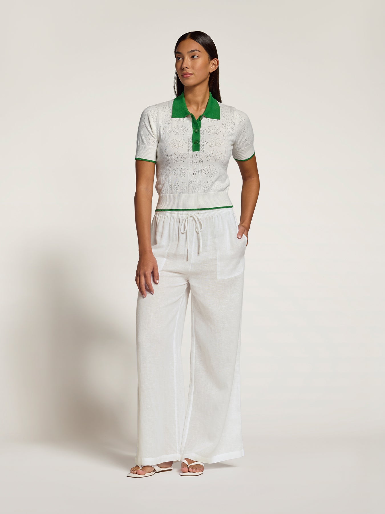 A woman modeling a white short-sleeve knit polo shirt with a green collar and green placket, paired with matching white wide-leg drawstring pants, against a plain off-white studio background.