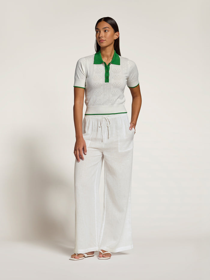 A woman modeling a white short-sleeve knit polo shirt with a green collar and green placket, paired with matching white wide-leg drawstring pants, against a plain off-white studio background.