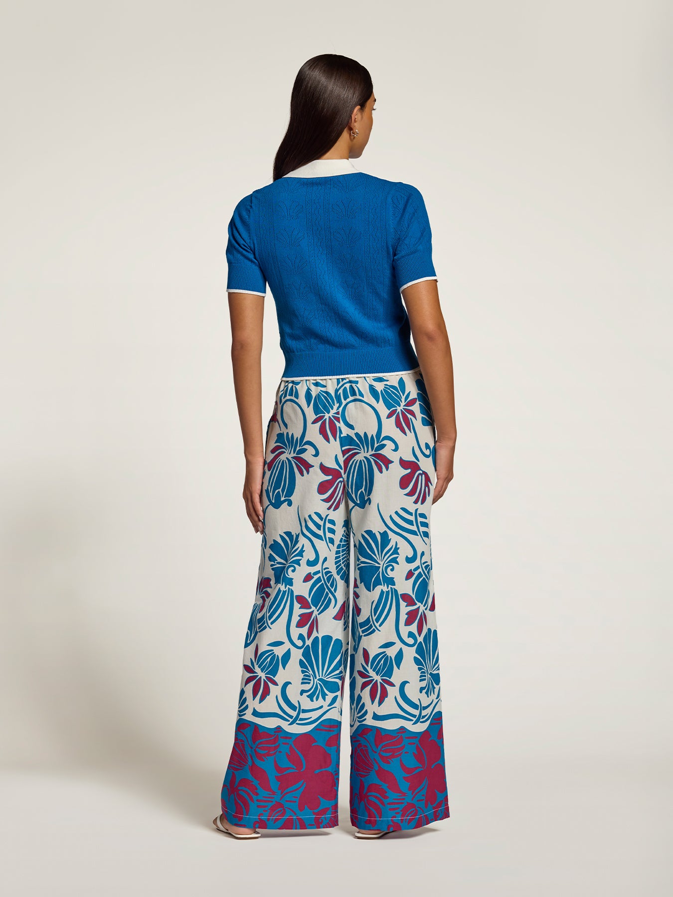 A model is shown from the back wearing a vibrant blue short-sleeve knit top and wide-leg trousers featuring a bold blue, magenta, and white floral pattern.