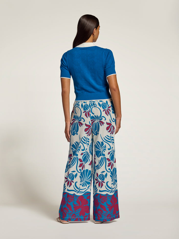 A model is shown from the back wearing a vibrant blue short-sleeve knit top and wide-leg trousers featuring a bold blue, magenta, and white floral pattern.