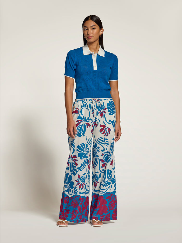 A model stands against a plain light beige studio background wearing a bright blue short-sleeve polo shirt with a white collar and cuffs, paired with wide-leg trousers featuring a bold blue, red, and white floral print.