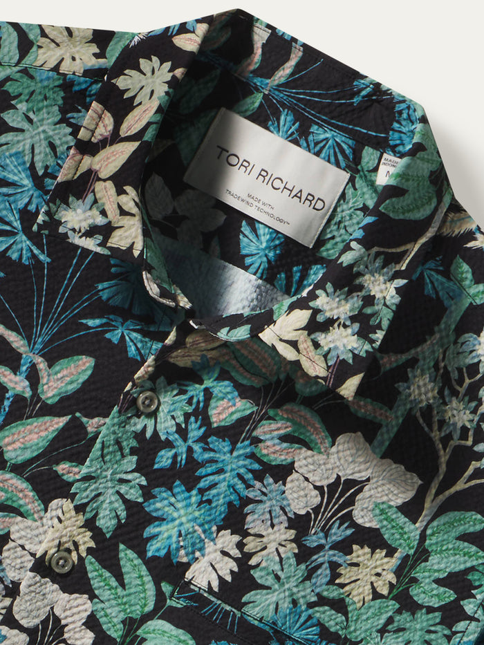 Close-up of a Tori Richard men's button-down shirt featuring a dense tropical floral and leaf print in teal, green, beige, and white on a black background.