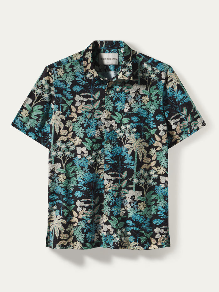 A short-sleeve button-up camp shirt with a dense tropical floral and palm tree print in teal, blue, beige, and black, displayed flat against a light off-white background.