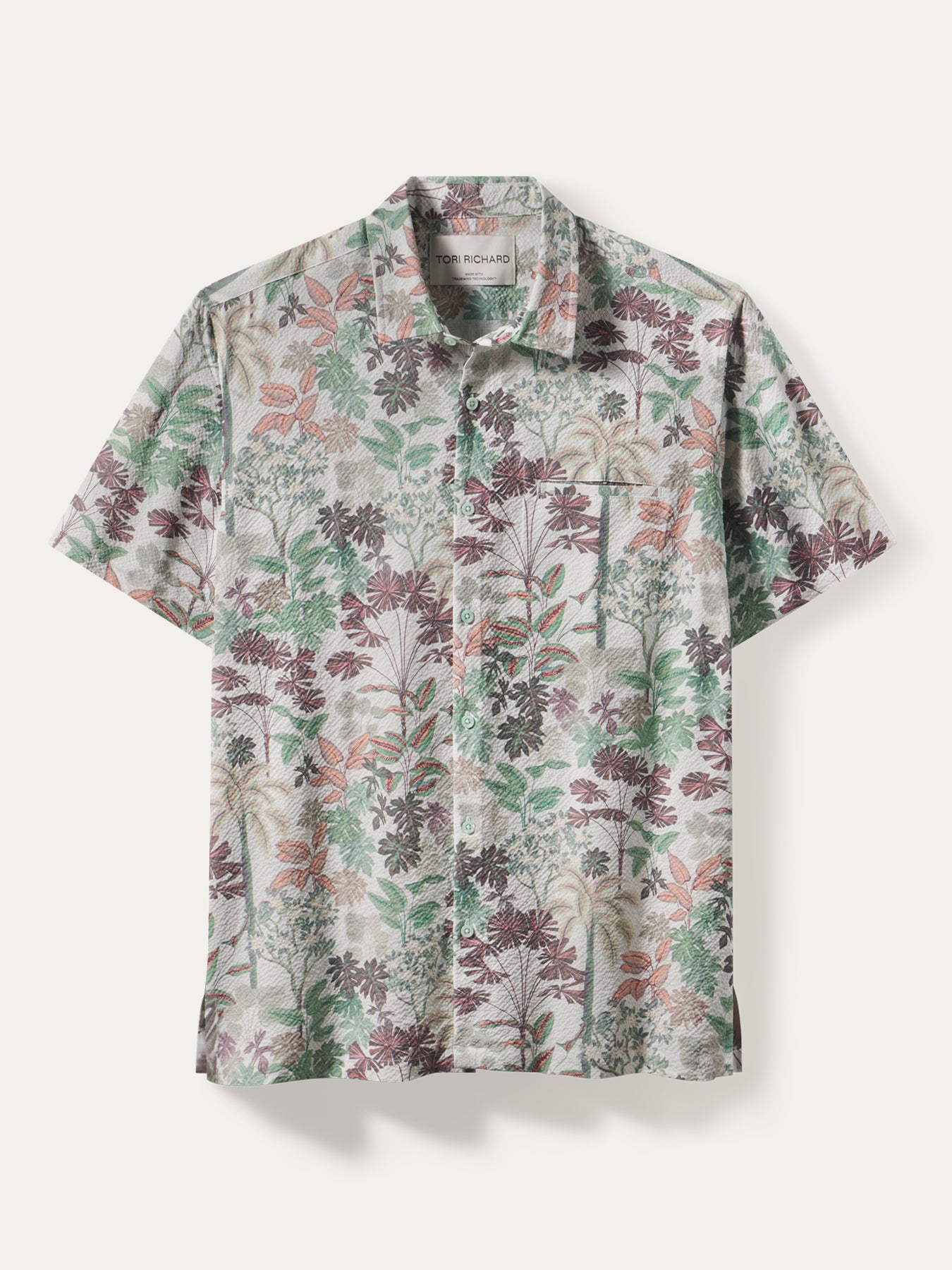 A short-sleeve button-up shirt with a dense botanical and floral print in muted green, dusty rose, and brown tones over a light beige base, displayed flat against a light off-white background.
