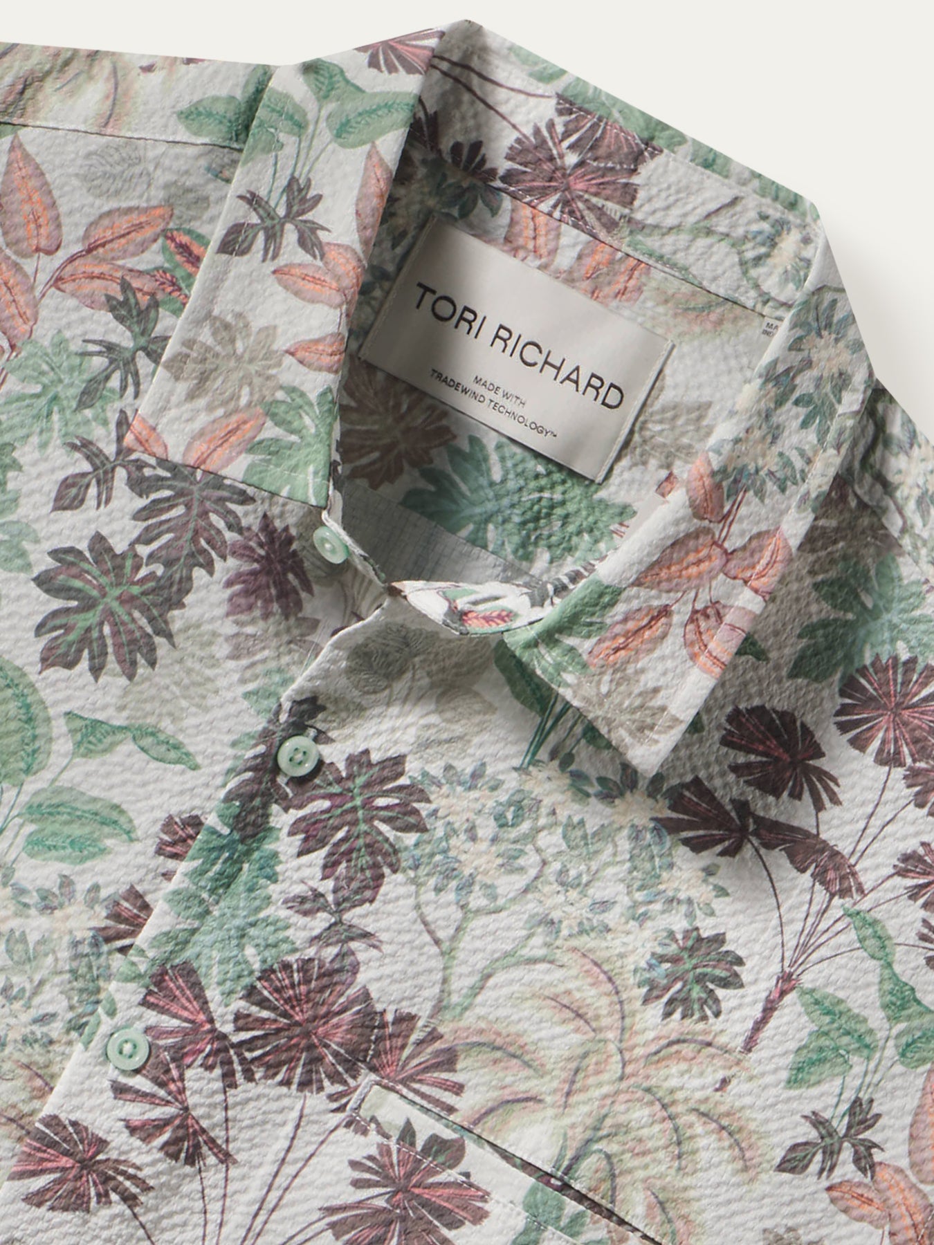 A close-up of a Tori Richard button-down shirt featuring a textured crinkle fabric and a botanical floral print in muted green, dusty rose, and brown tones on a light cream background.