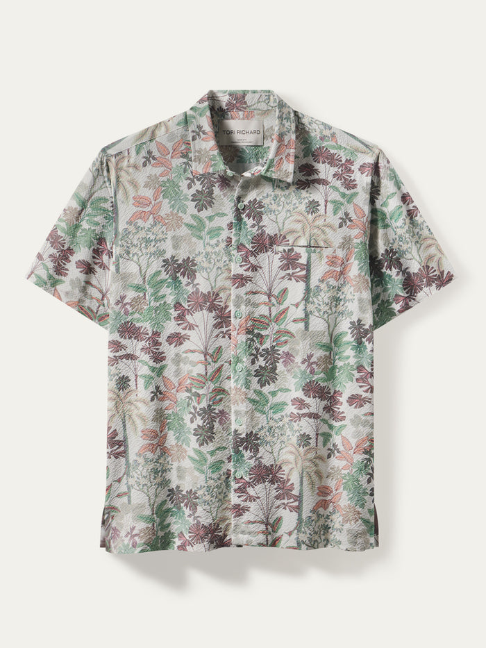 A short-sleeve button-up shirt with a dense botanical and floral print in muted green, dusty rose, and brown tones over a light beige base, displayed flat against a light off-white background.