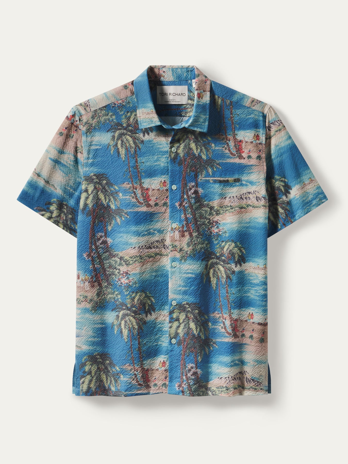 A short-sleeve button-up camp shirt with a vibrant blue tropical beach print featuring palm trees, sandy beaches, and water, displayed flat against a light beige background.