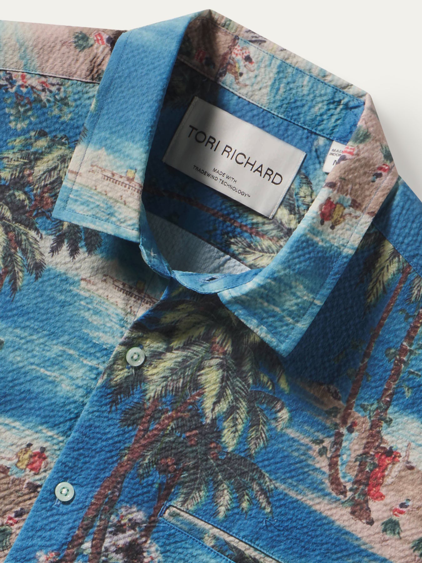 Close-up of a collared button-up shirt by Tori Richard, featuring a blue tropical palm tree print with a textured finish and a white brand label visible inside the collar.