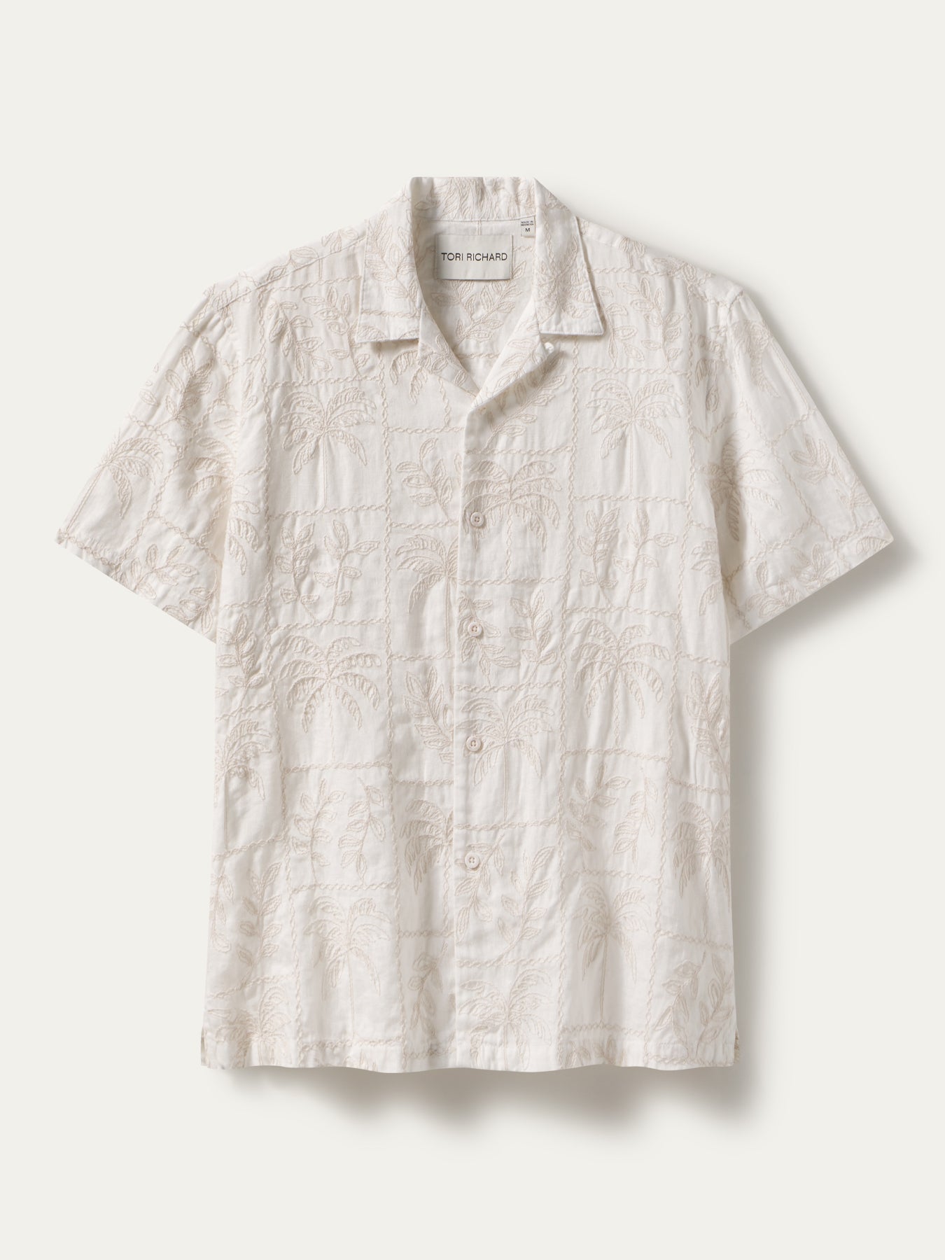A short-sleeve camp collar button-up shirt in a warm off-white or cream color, featuring an all-over tonal embroidered pattern of palm trees and geometric lines. The shirt is displayed flat against a light beige background.
