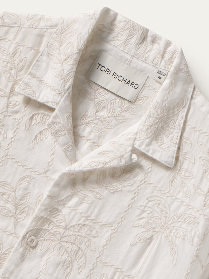 Close-up of a white cotton shirt by Tori Richard featuring a delicate beige floral and leaf embroidery pattern, with the brand label visible inside the collar.