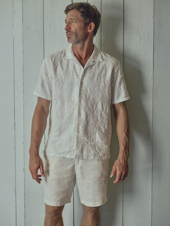 A man wearing a white short-sleeve button-up shirt with a subtle textured pattern and matching white linen shorts, standing against a white wooden plank wall.
